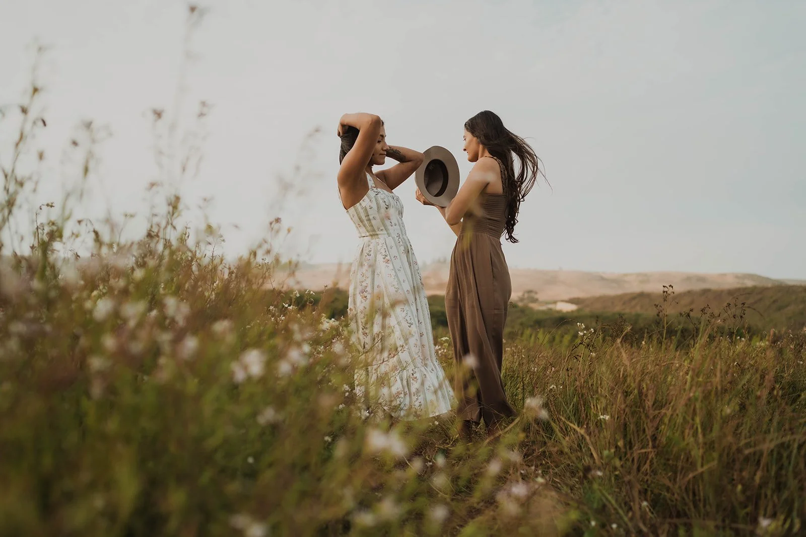 Two women stand in a grassy field during sunset, one holding a hat while the other adjusts her hair, both wearing summer dresses.