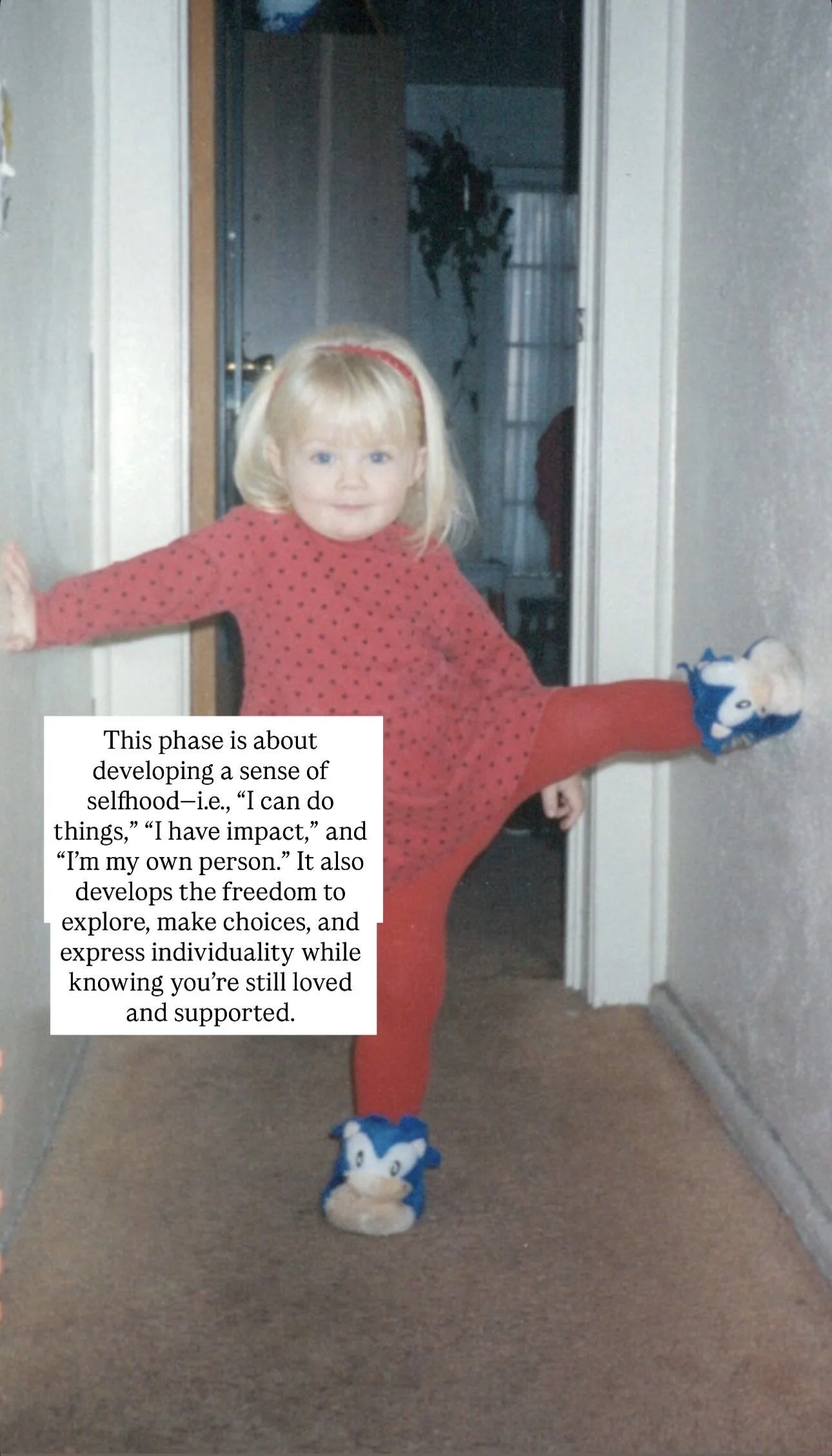 A young girl with blond hair is stretching her leg and arm, wearing red polka dot pajamas and Sonic the Hedgehog slippers, standing in a hallway at home.