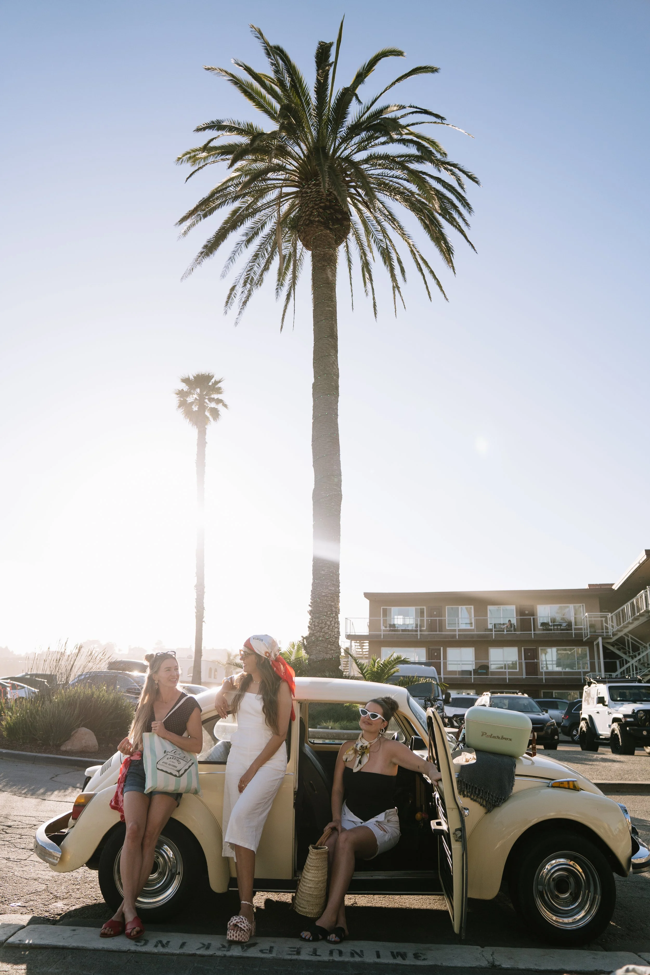 Three women in summer clothing and sunglasses standing and sitting around a vintage beige Volkswagen Beetle parked in a lot near palm trees and an apartment building, with clear blue sky and bright sunlight.