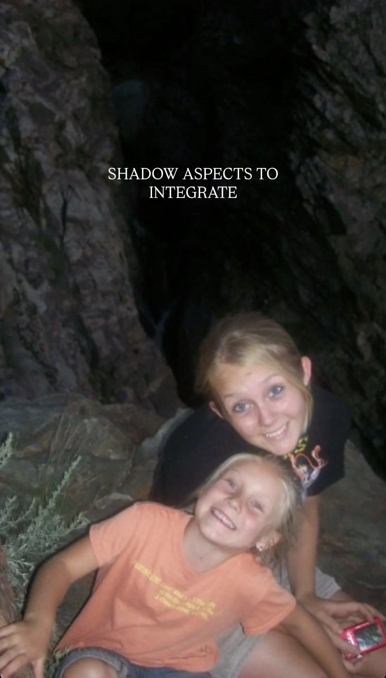 Two children, a girl with blonde hair and a girl with light brown hair, are smiling and looking up at the camera inside a rocky crevice.