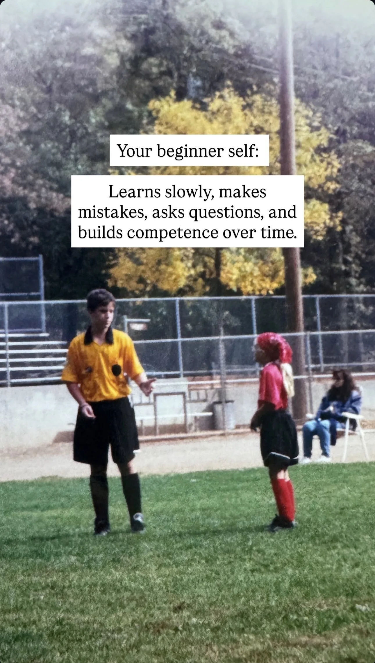 A referee in a yellow shirt and black shorts talking to a young female soccer player with a red wig and red socks on a green field, with spectators in the background.