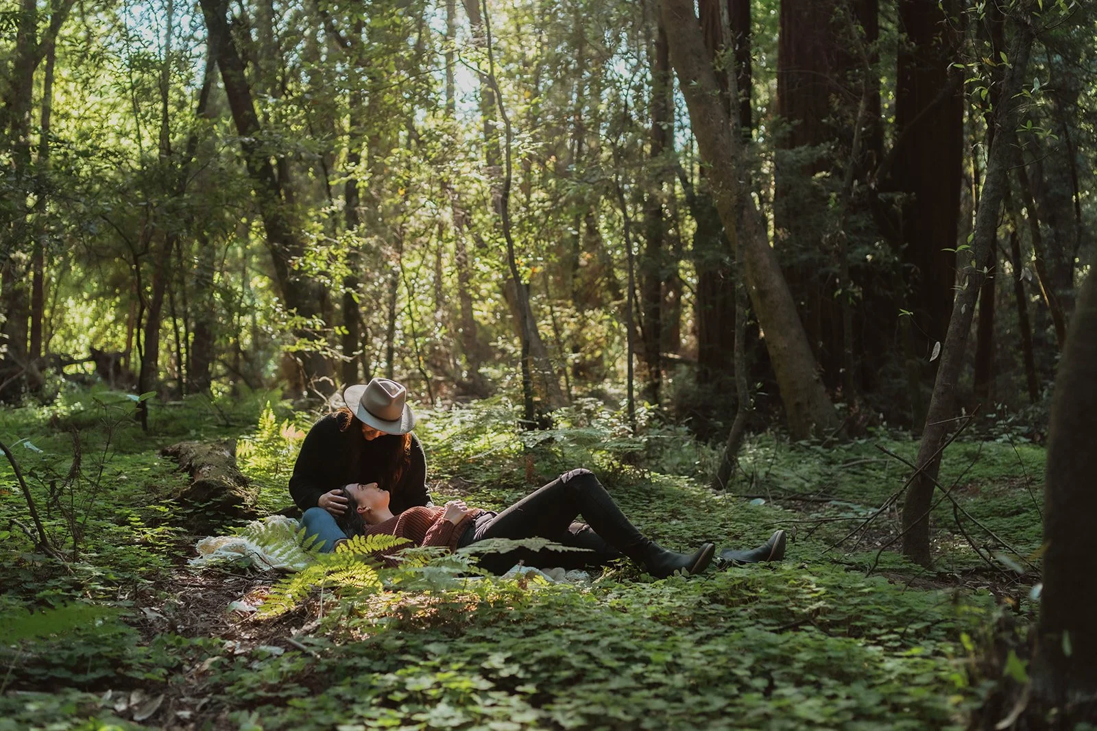 A woman lying down on a blanket in a forest, being cared for by another woman wearing a hat, surrounded by tall trees and greenery.
