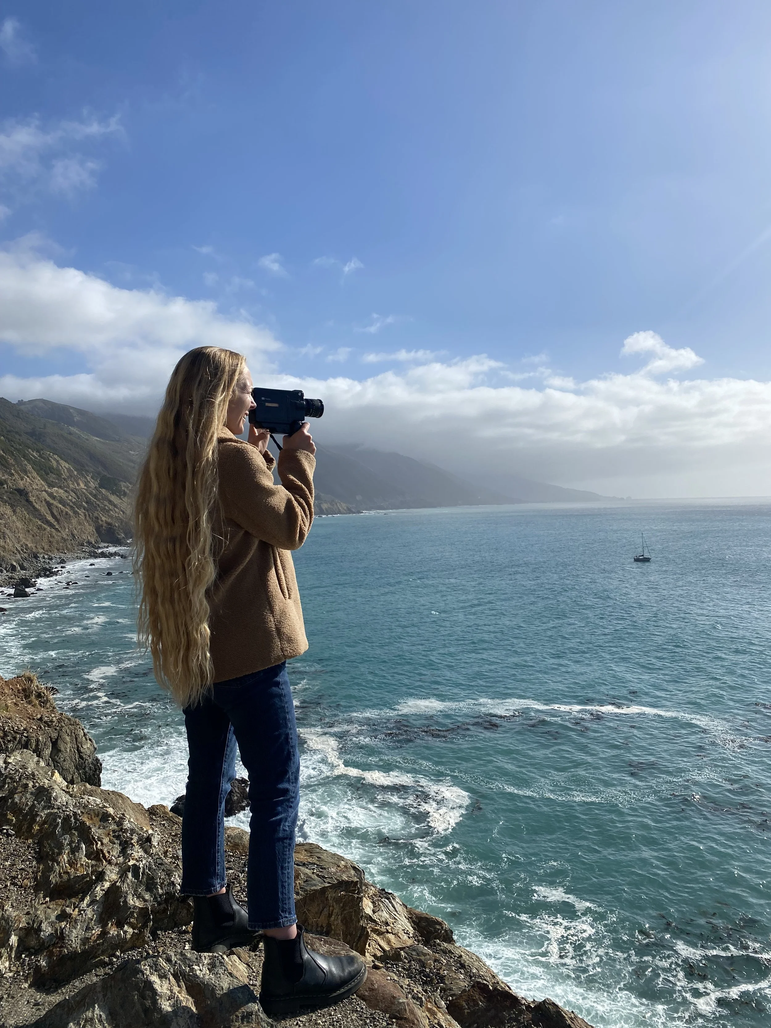 A woman with long blonde hair, dressed in a brown jacket, jeans, and black boots, standing on rocks by the ocean, taking a photo with a camera during daytime with clear blue skies and distant mountains.