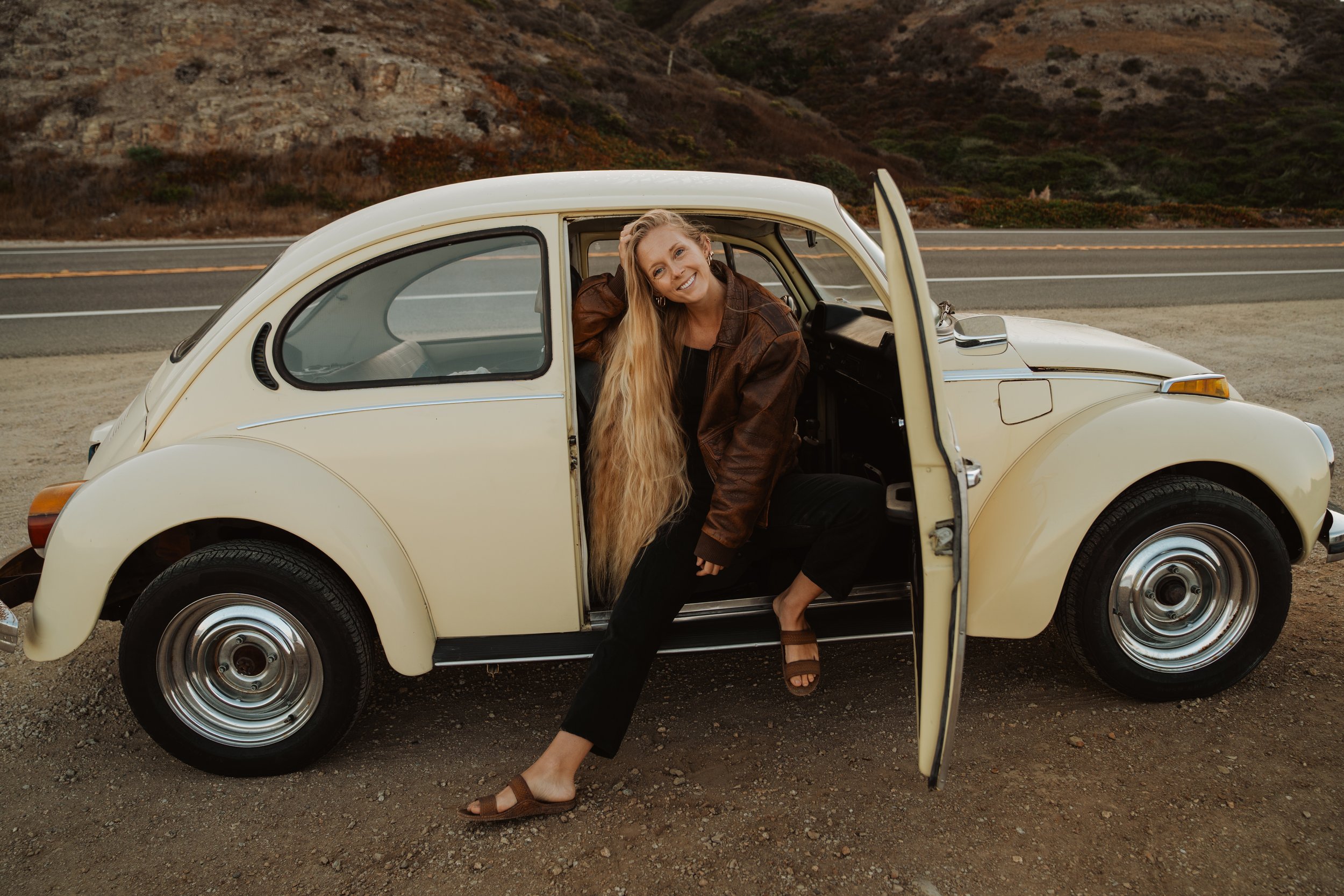 A woman with long blond hair sitting in an open door of a vintage cream-colored Volkswagen Beetle parked on the side of a road with desert landscape and hills in the background.