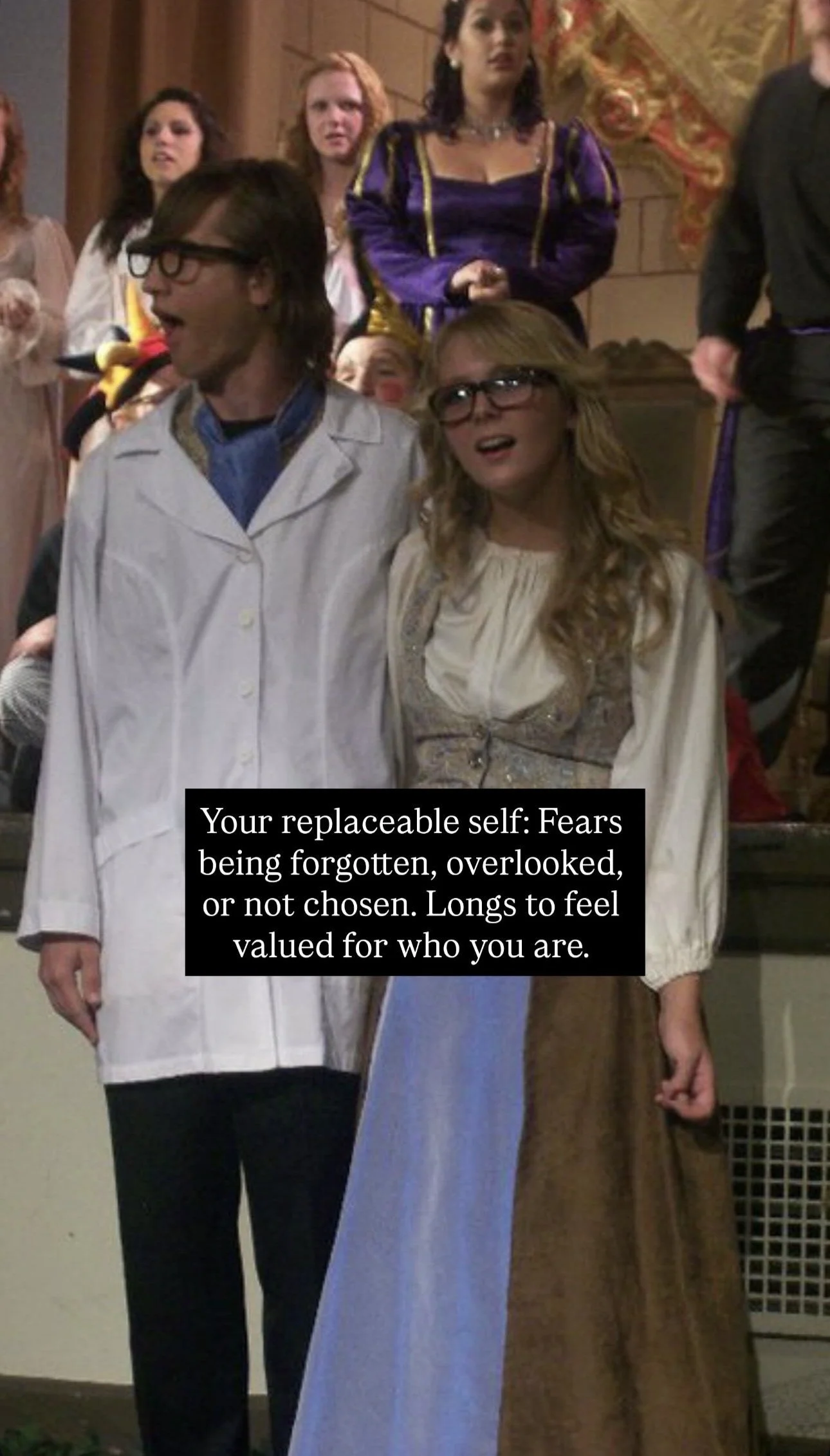 Scene from a theatrical performance showing two actors in the foreground, one in a white lab coat and glasses, the other in a long dress and glasses. Behind them, a group of people stand, some dressed in costumes, including a woman in a purple and ye