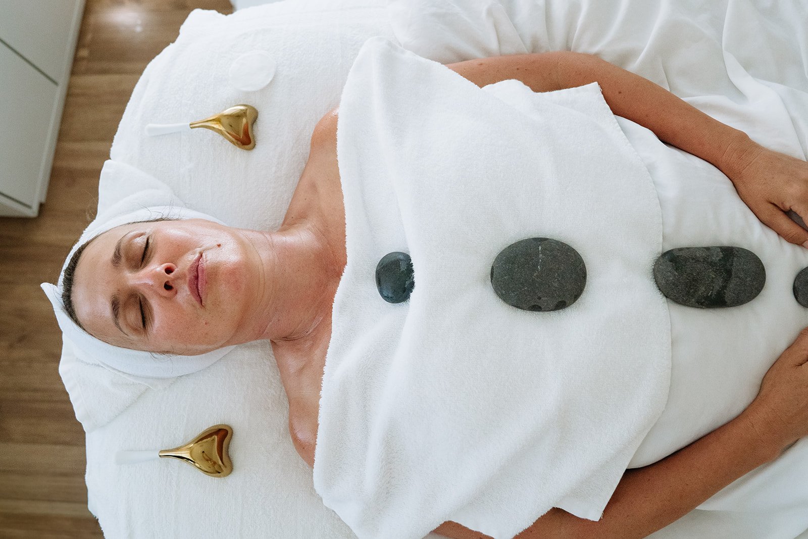 A woman lying on a spa bed with black massage stones on her abdomen, a white towel covering her chest, and two gold decorative objects near her head, in a relaxing spa setting.