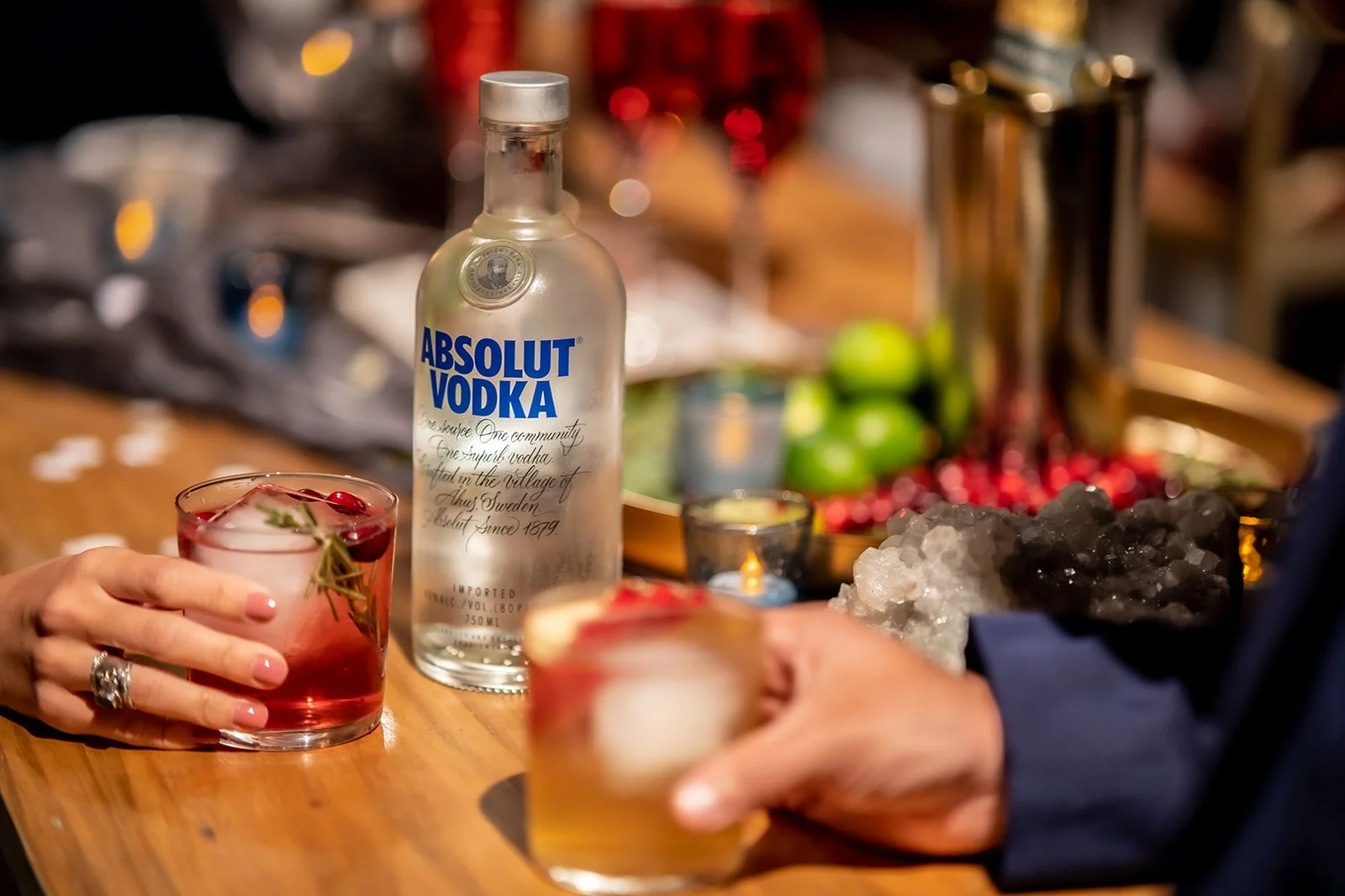 A table with a bottle of Absolut Vodka, a pink cocktail with cherries and herbs, and a drink with ice, with hands holding the cocktails, and a background with liquor bottles, limes, and ice.