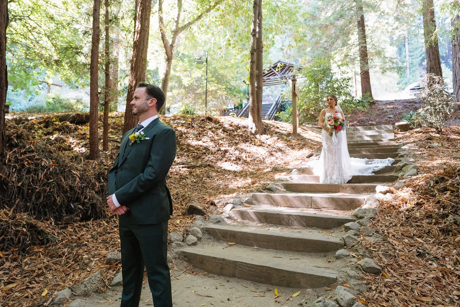 A bride holding a bouquet stands on stone steps in a wooded outdoor area, with a groom in a dark suit standing nearby, both present at a wedding ceremony.