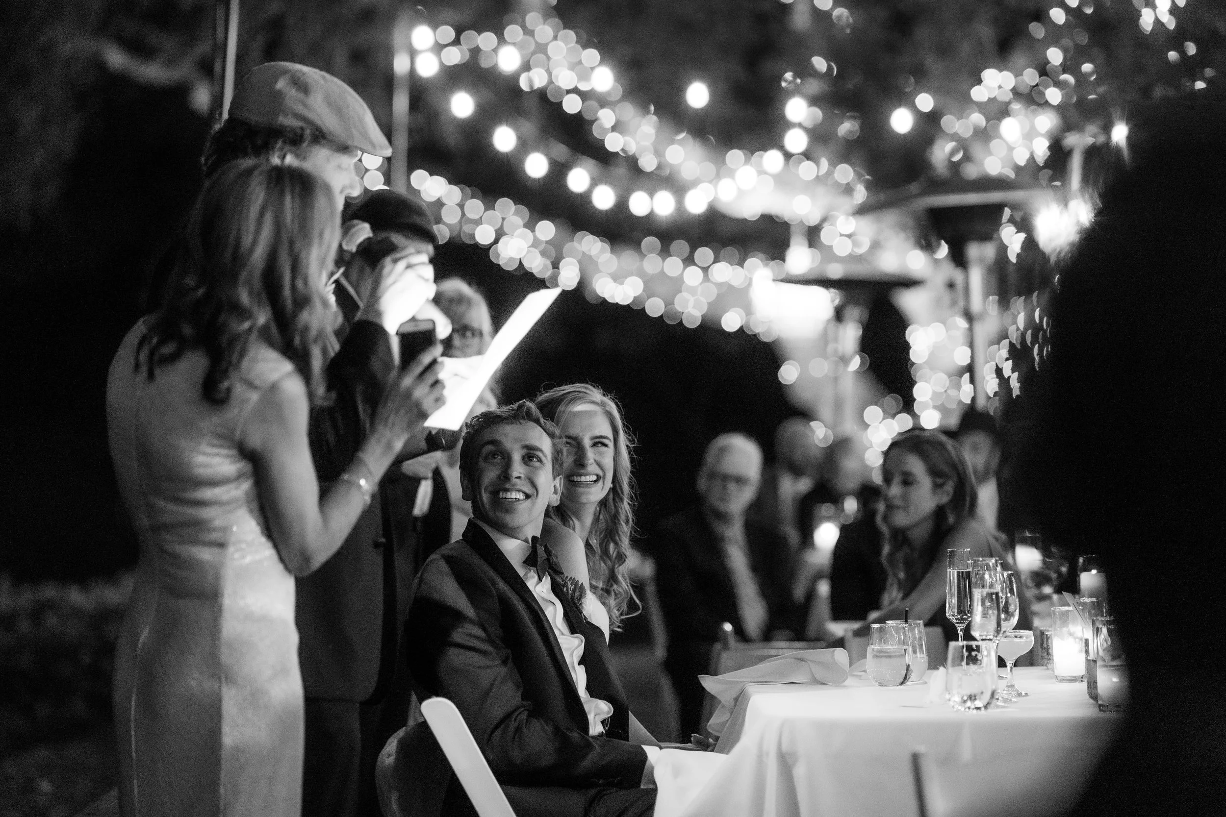 Black and white photo of a wedding celebration with the bride and groom sitting at a table, surrounded by friends and family, with string lights hanging overhead.