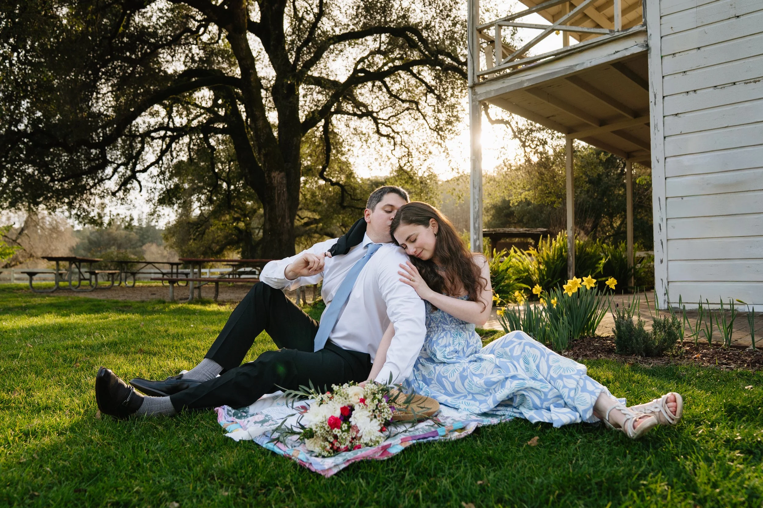 A young couple sitting on a blanket in a park during sunset, embracing tenderly with a bouquet of flowers nearby.