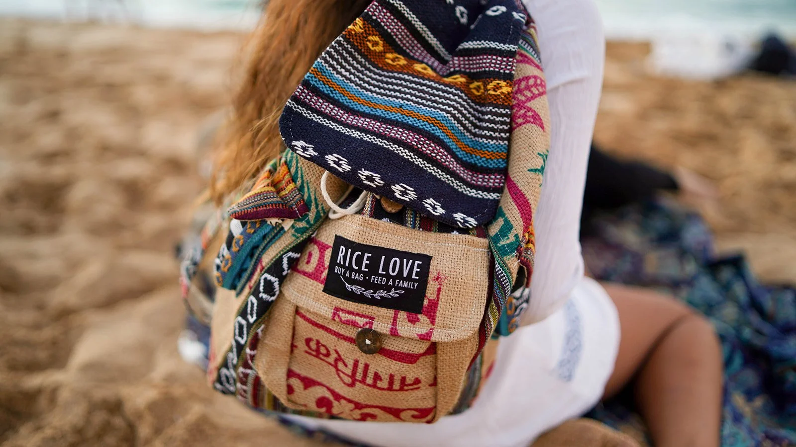 Close-up of a person on a beach wearing a patchwork bag with a label 'RICE LOVE Buy a bag, feed a family' and colorful woven textiles. The background shows sand and a blurred ocean.