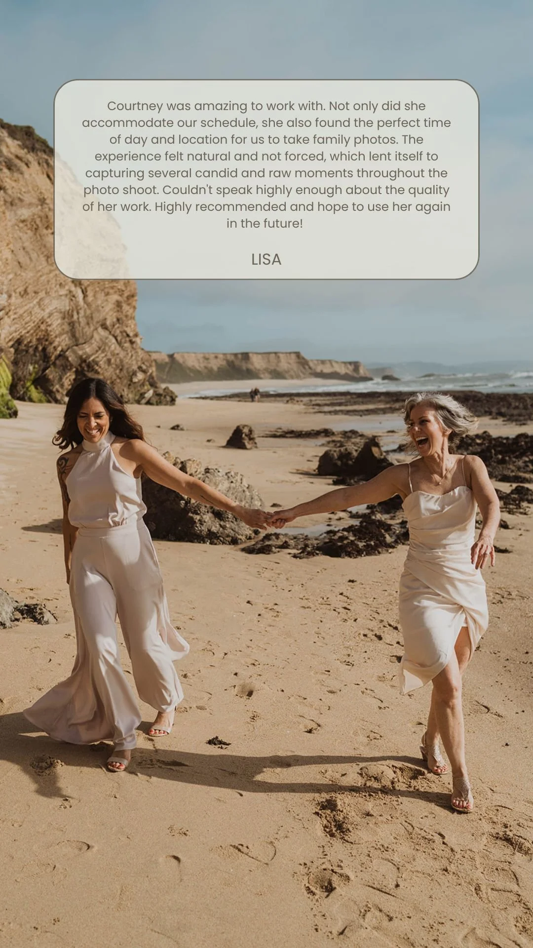 Two women in white dresses holding hands and running on a sandy beach with rocks, cliffs, and ocean in the background.