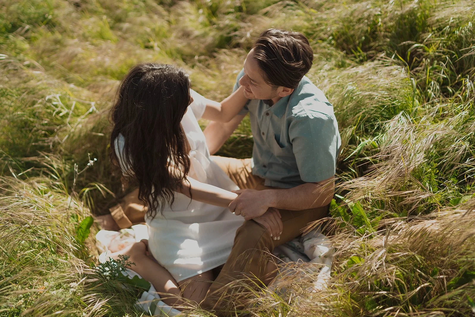 A couple sitting in a field of tall grass, smiling and engaging in a close, intimate moment.