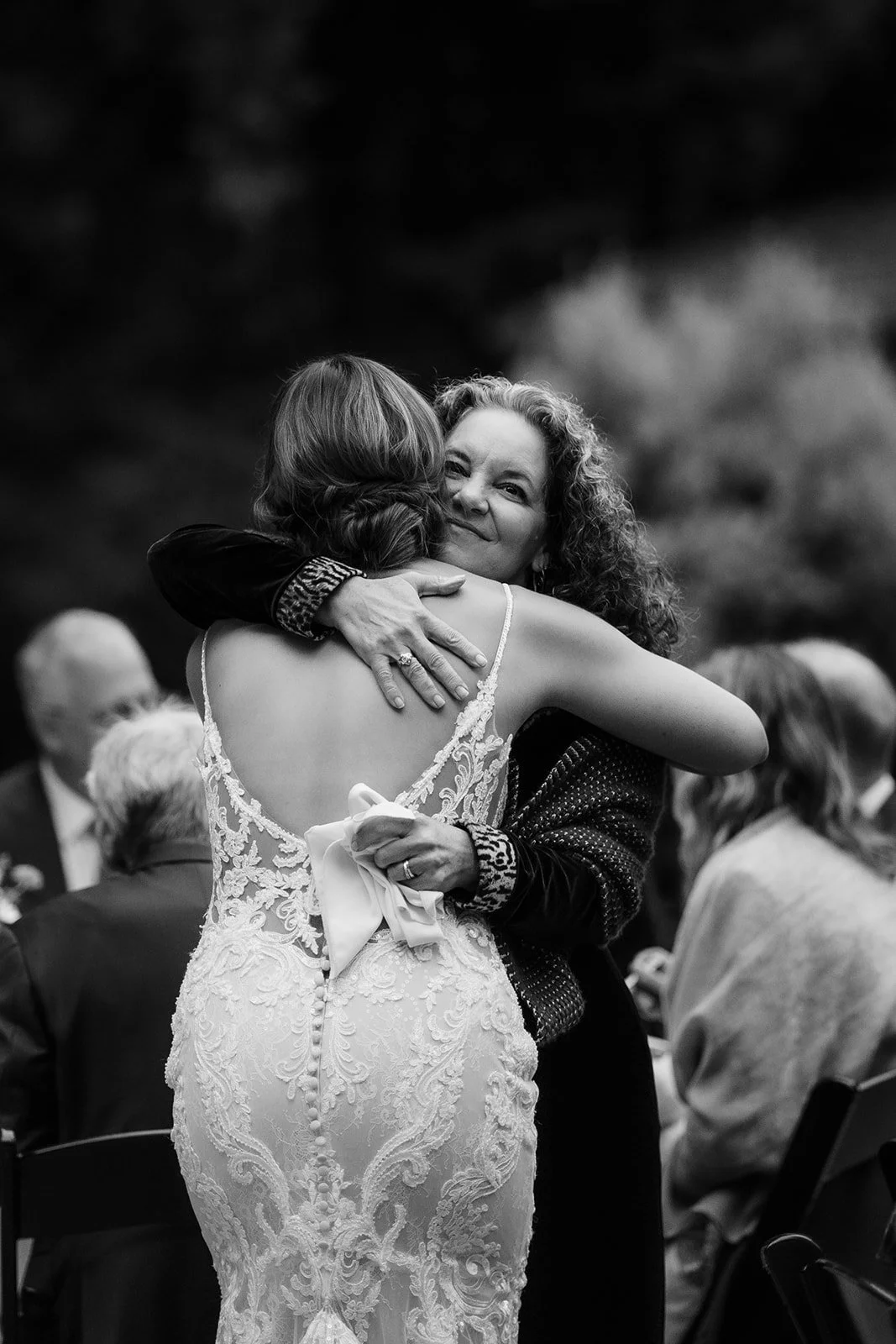 A woman in a lace dress and a woman in a dark jacket hugging at a wedding reception, with other guests seated in the background.
