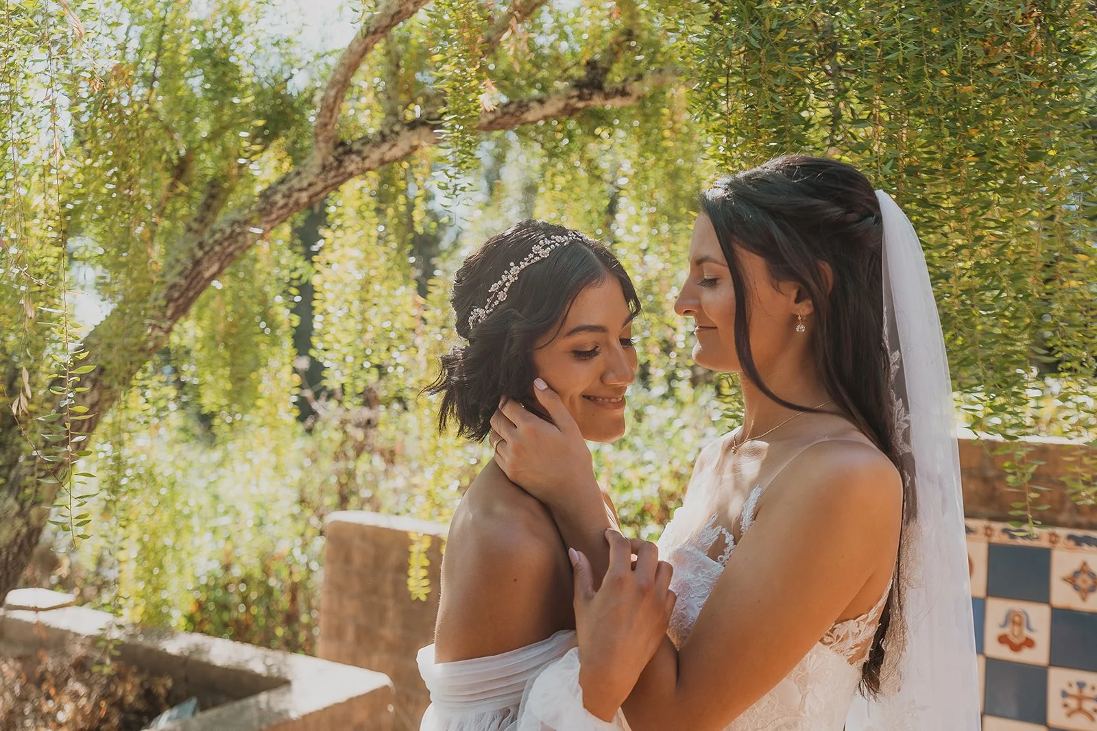 Two women, one in a wedding dress and veil, and the other in a white dress, embrace outdoors among green trees and leaves, smiling with eyes closed.