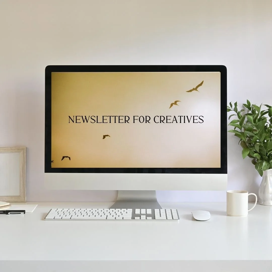Computer monitor displaying 'Newsletter for Creatives' with a beige background and flying bird graphics on a white desk with a plant, mug, frame, and keyboard.