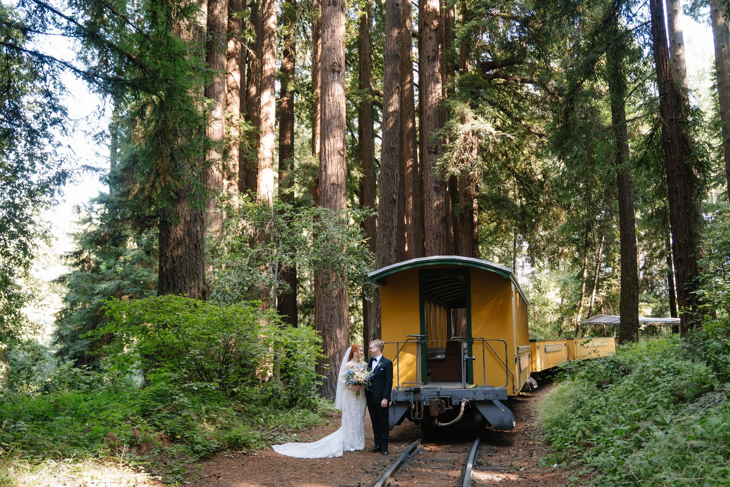 A bride and groom standing on train tracks in a forest, leaning toward each other, with the bride holding a bouquet.