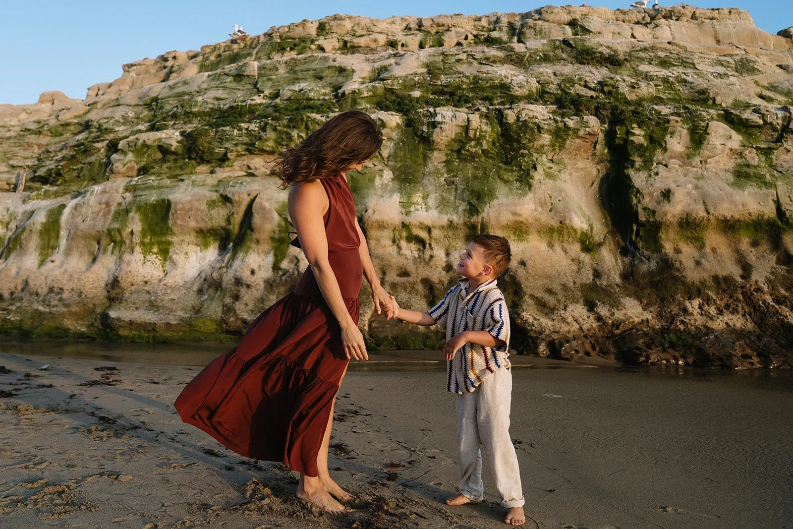 A woman and young boy holding hands on a beach with wet sand and a large mossy rock formation in the background at sunset.