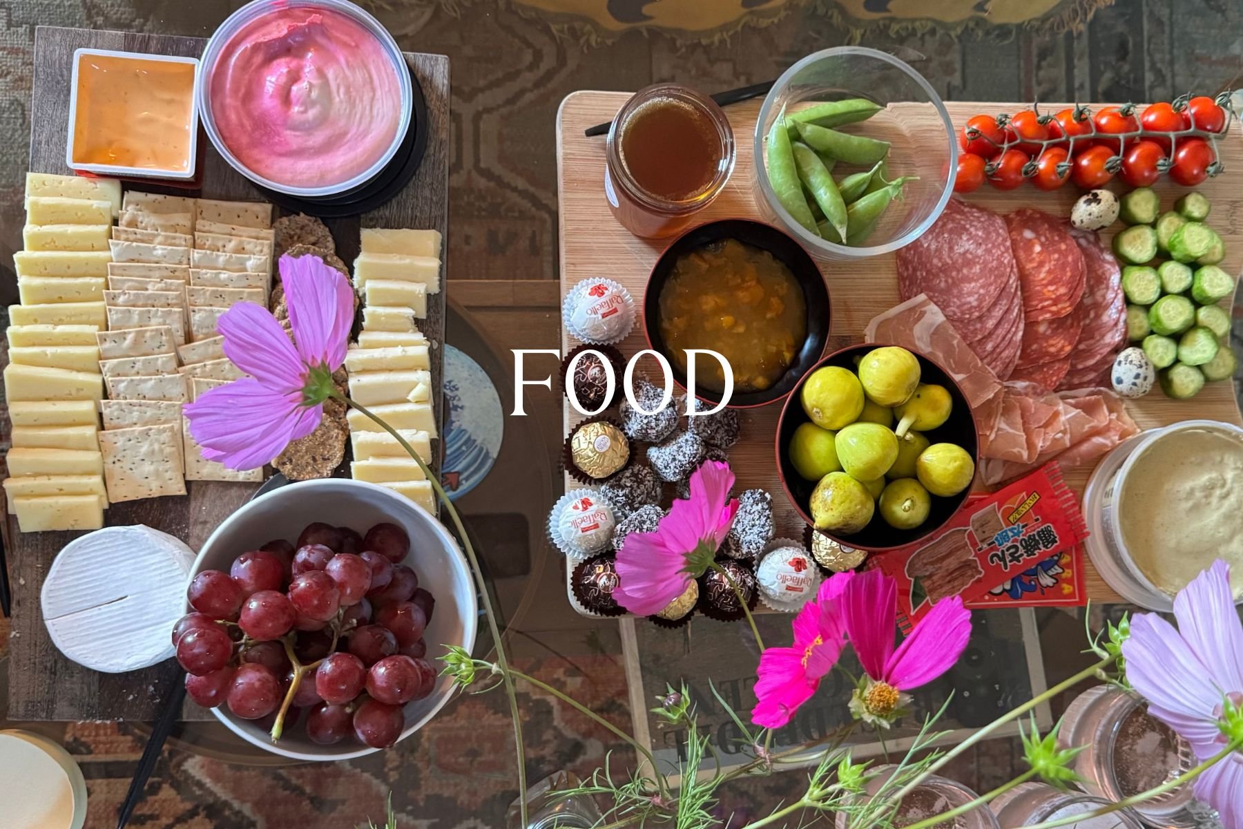 Assorted cheese, fruits, vegetables, crackers, and chocolates on a table, decorated with pink flowers.