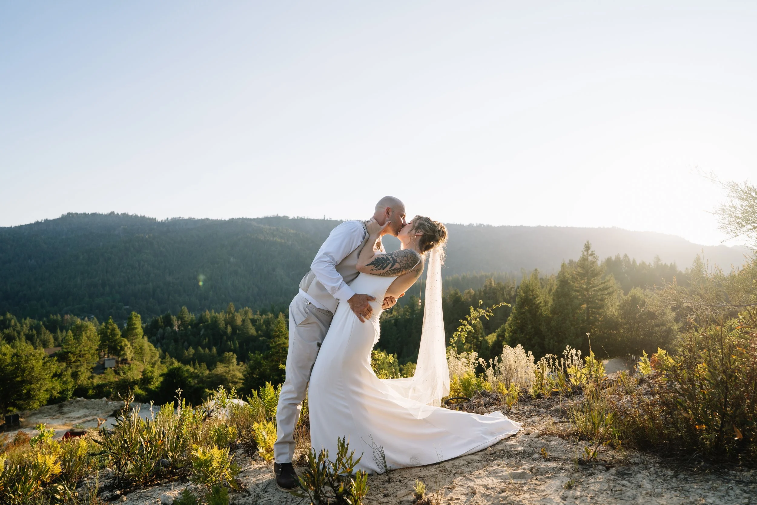 A couple in wedding attire sharing a kiss outdoors on a sunny day, with mountains and trees in the background.