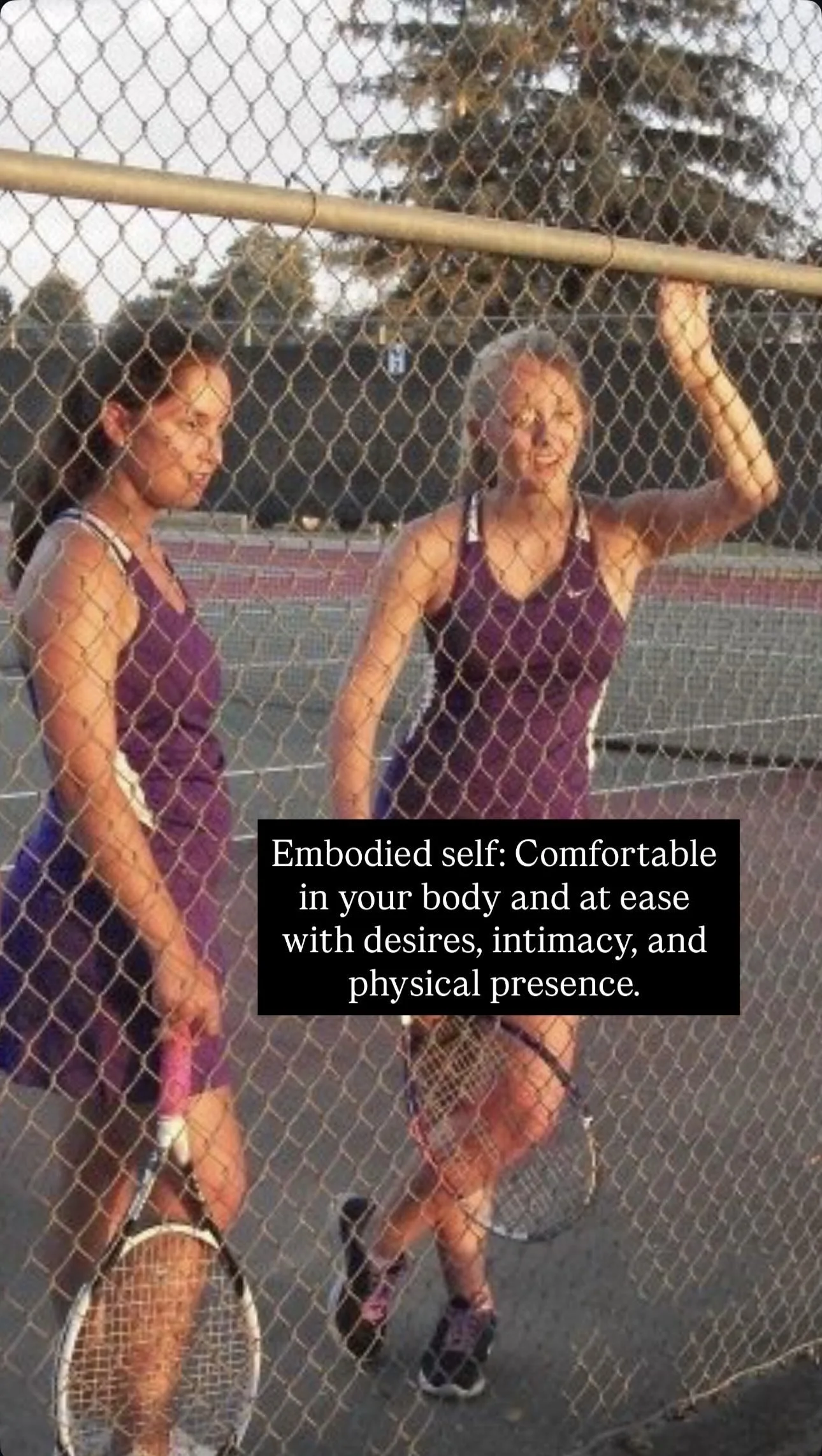 Two women in tennis outfits standing behind a chain-link fence, holding tennis rackets during sunset at an outdoor tennis court.