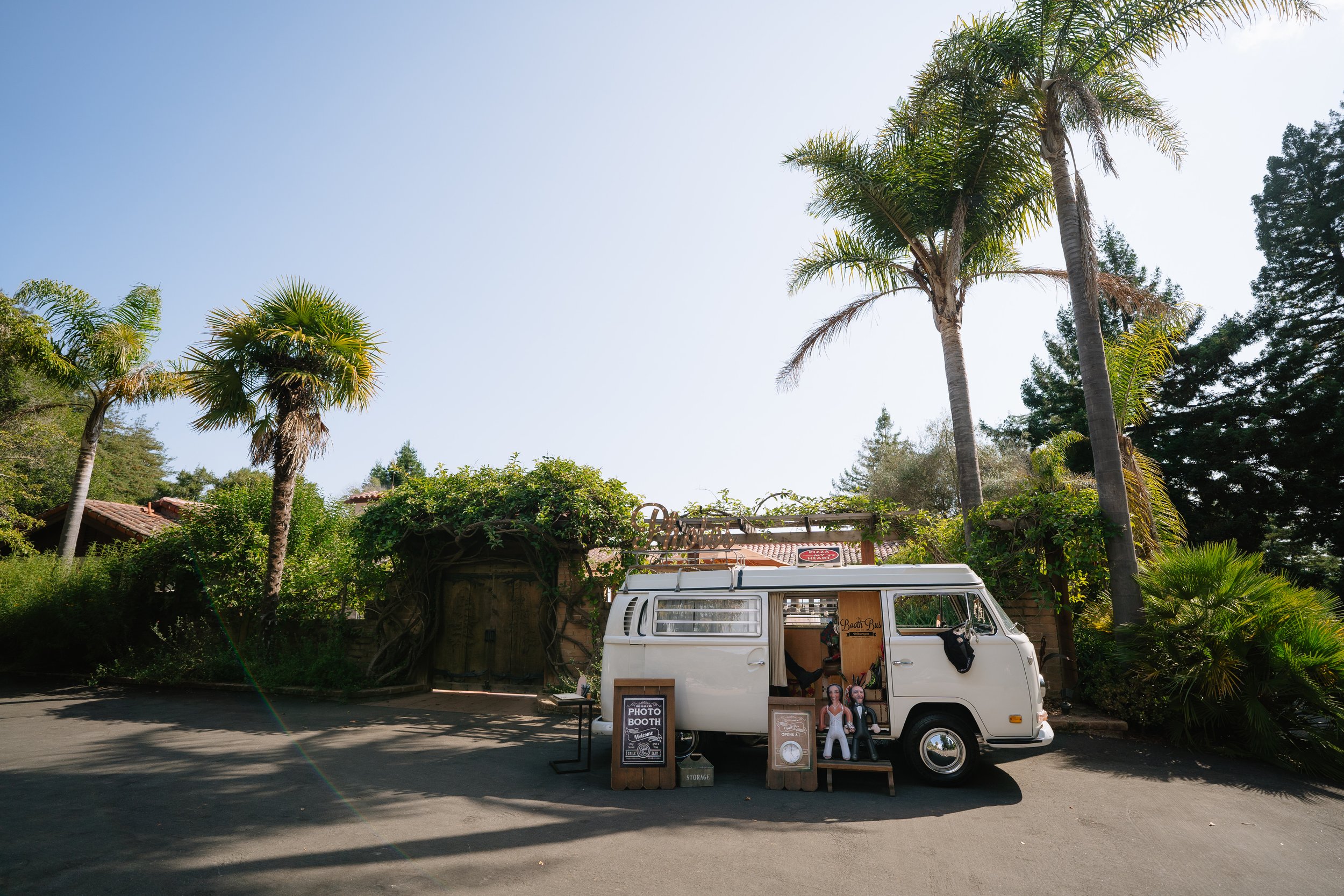 A vintage white van parked outdoors with a small photo booth setup, various signs and decorations, surrounded by palm trees and greenery on a sunny day.