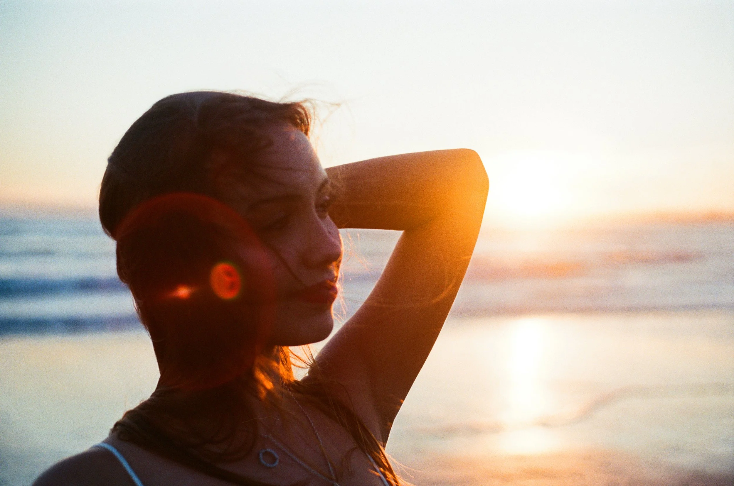 A woman with wet hair standing at the beach during sunset, with her eyes closed and one arm raised behind her head.