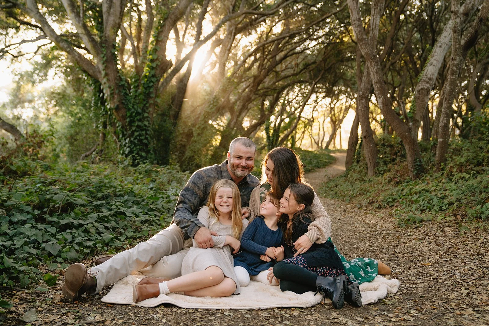 A family of five, including two adults and three children, sitting on a blanket on a dirt path in a wooded area during sunset, enjoying each other's company.