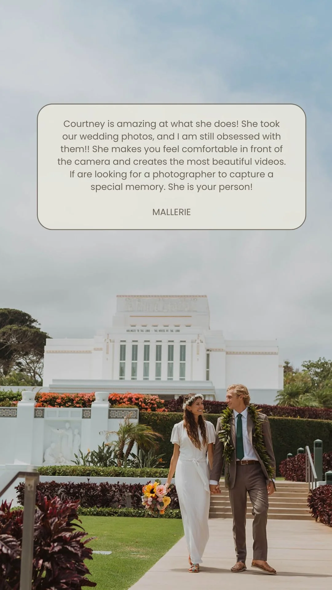 A newlywed couple walking hand in hand outdoors, with a white historic building in the background, surrounded by greenery and flowers, during a wedding photoshoot.