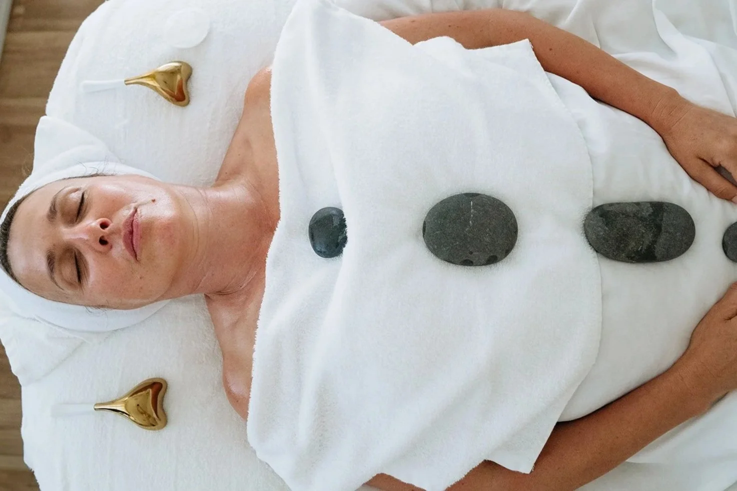A woman lying on a spa bed with hot stones on her chest, a towel covering her torso, and a white headband around her head.