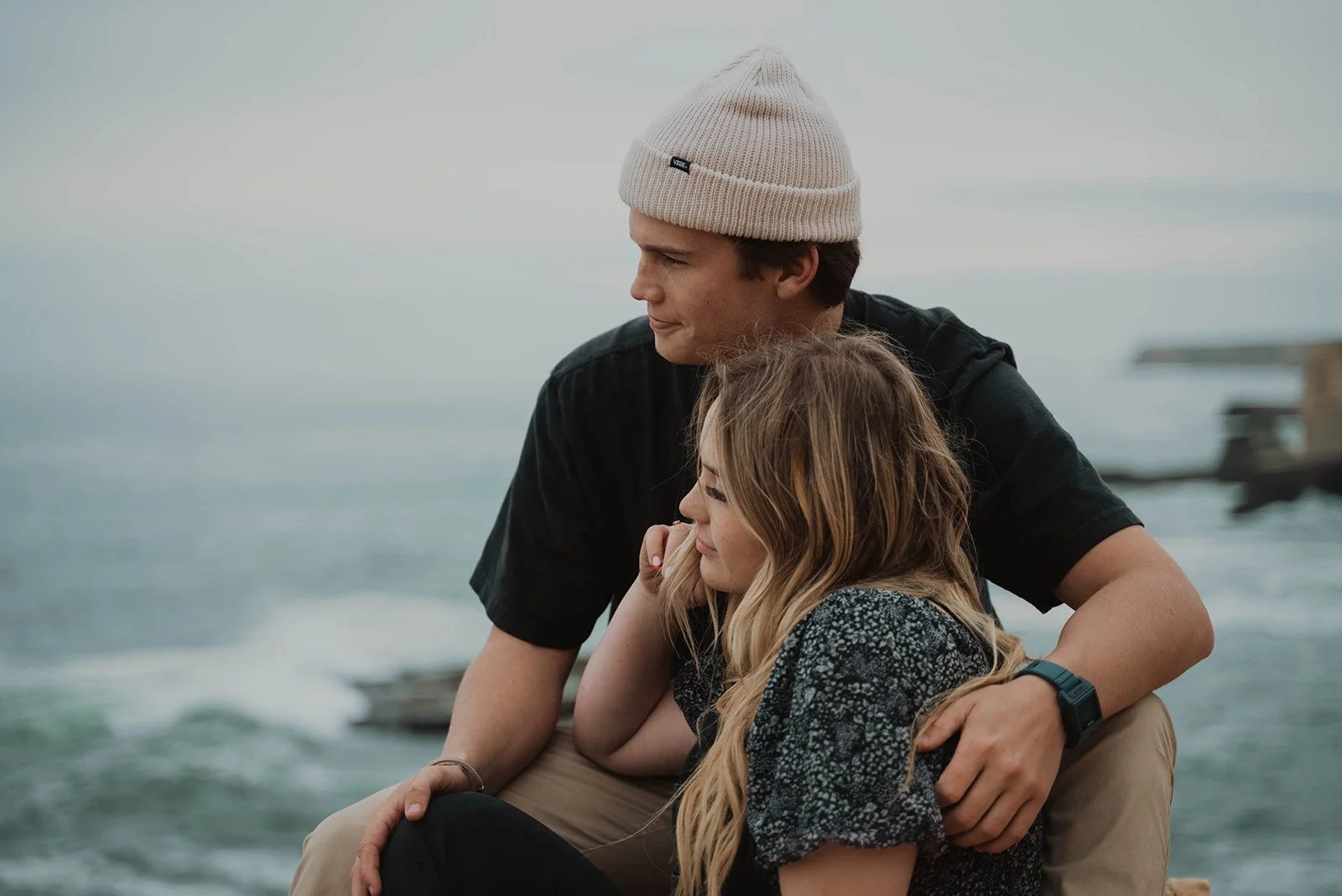 A young couple sitting by the seaside, embracing and gazing at the ocean. The man is wearing a beige knitted beanie and black shirt, while the woman has long blonde hair and a patterned top.