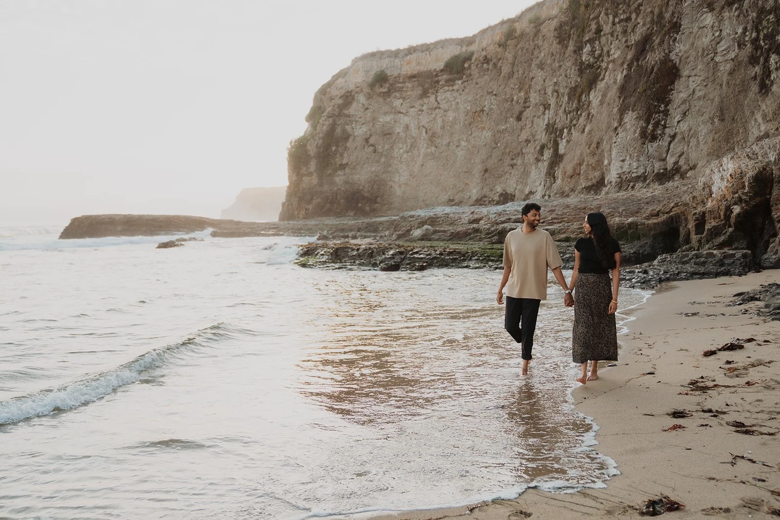 A couple walking hand in hand along the shoreline of a beach with cliffs in the background.