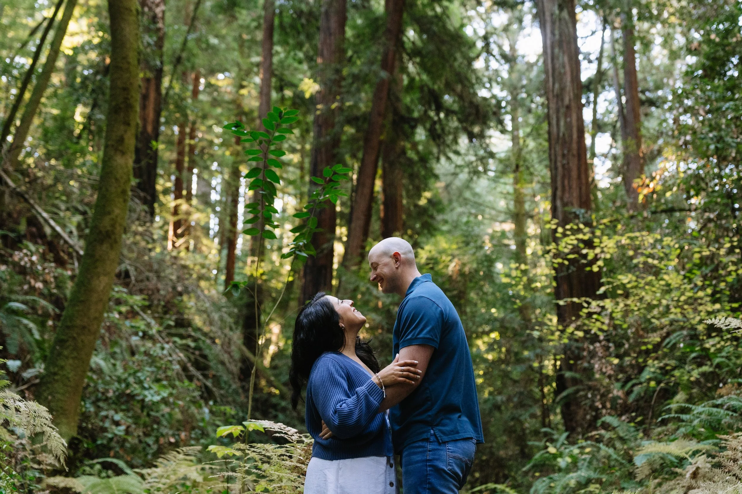 A couple smiling and looking at each other in a forest with tall trees and green foliage.