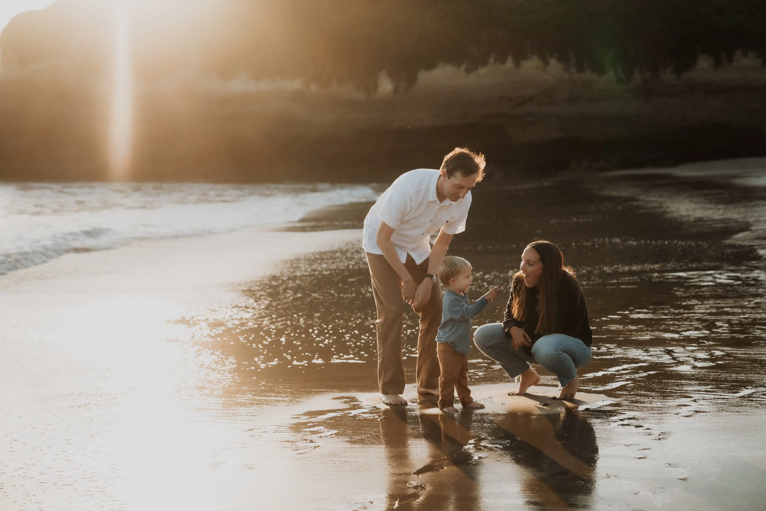 A family of three enjoying time together on the beach at sunset, with a man, woman, and small child.