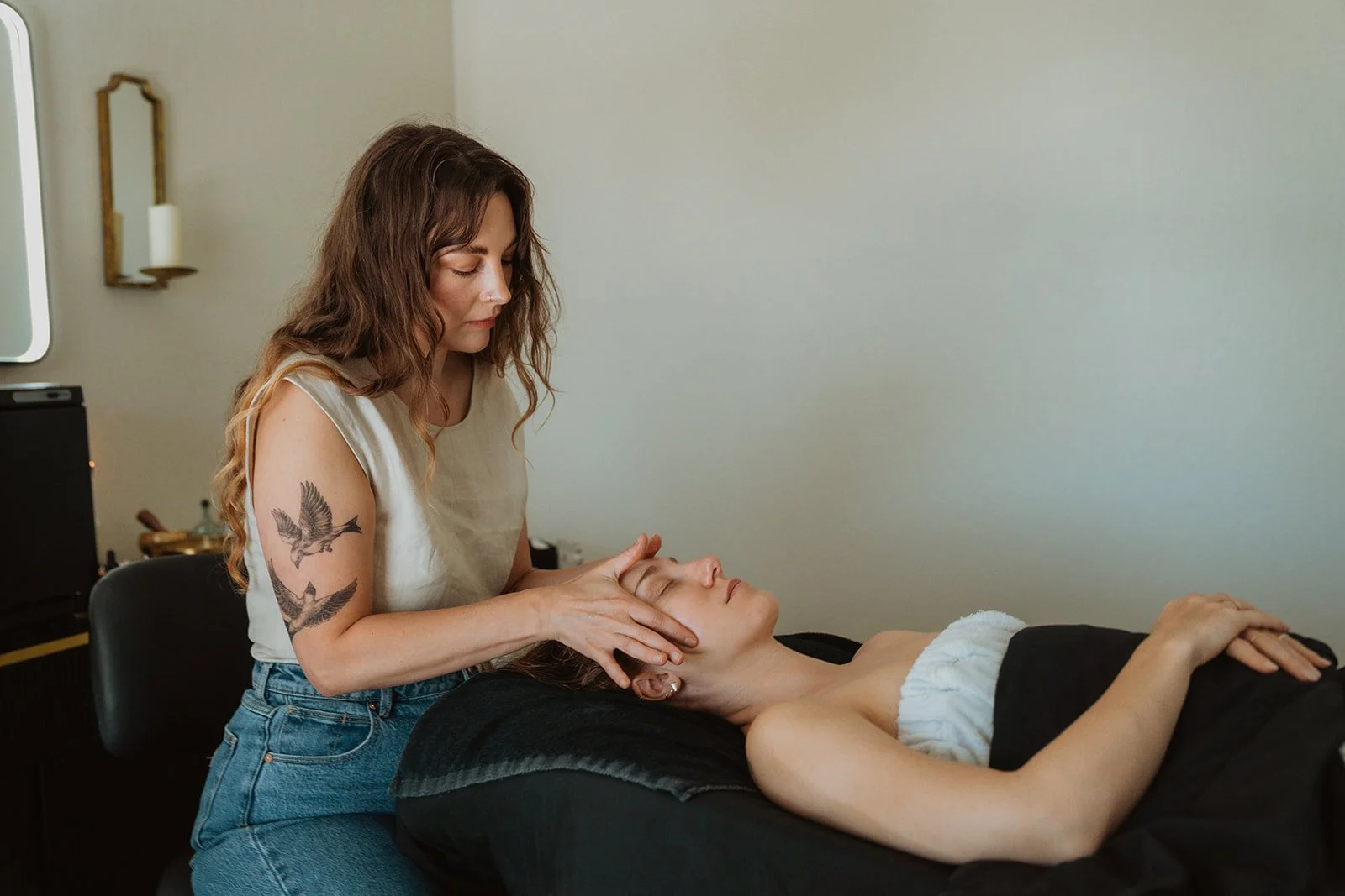 A woman giving a facial massage to another woman who is lying on a treatment bed with a black towel, in a calming room.