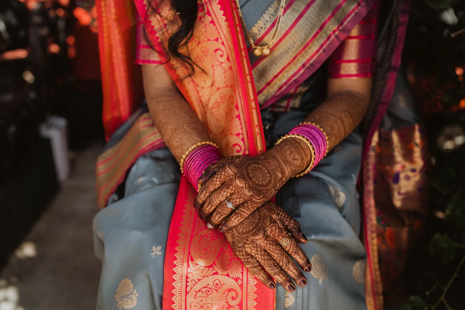 Close-up of a person wearing traditional Indian attire, showing decorated henna-adorned hands and wrists with pink bangles, sitting with hands folded in lap.