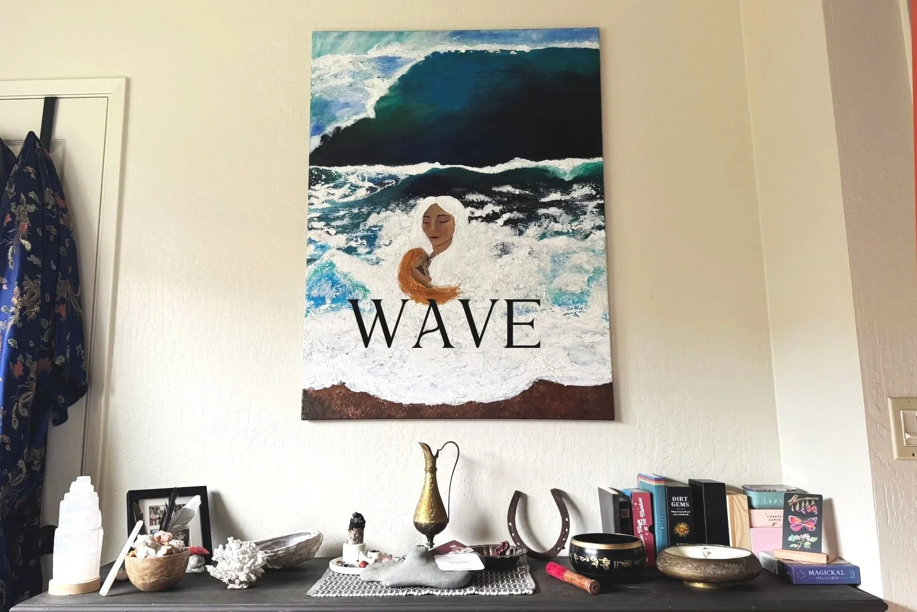 A painting of a woman with long hair in the surf with the word 'WAVE' written on it, hung on a wall above a cluttered table with books, a horseshoe, bowls, and decorative objects