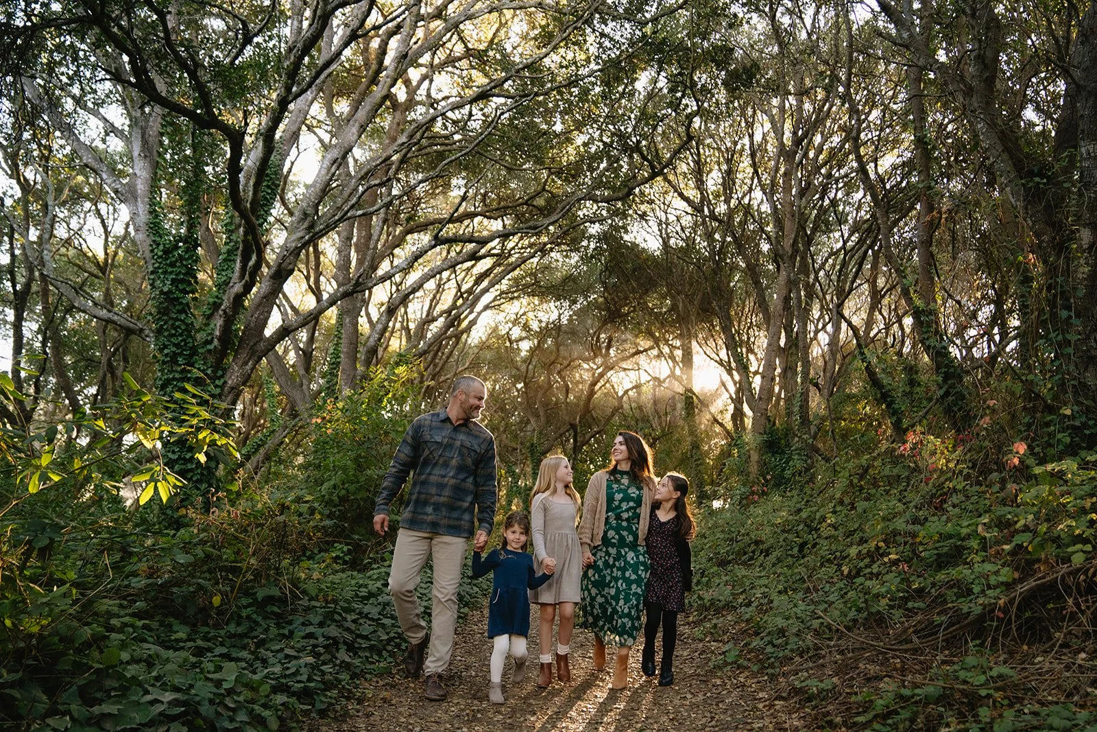 A family of six walking along a trail in a wooded forest during the sunset, smiling and holding hands.