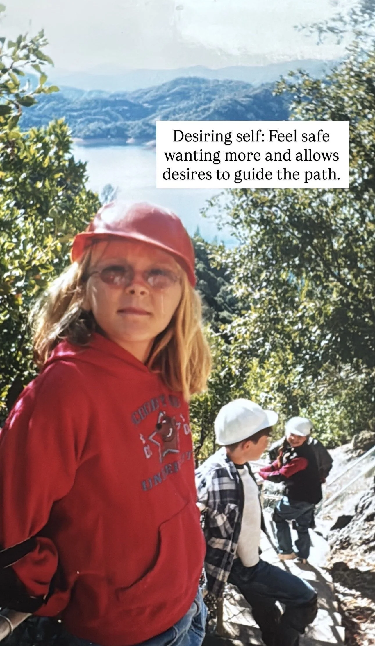 A girl in a red hoodie and red cap stands outdoors with mountains and trees in the background, with two boys in the background near rocks, all wearing casual outdoor clothing.