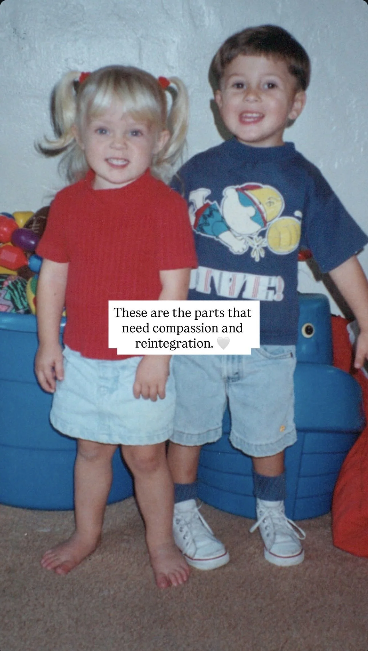 A young girl with blonde hair in pigtails, wearing a red shirt and light blue shorts, standing next to a boy with brown hair, wearing a navy blue T-shirt and light blue shorts, in a room with stuffed toys and a blue storage bin in the background.