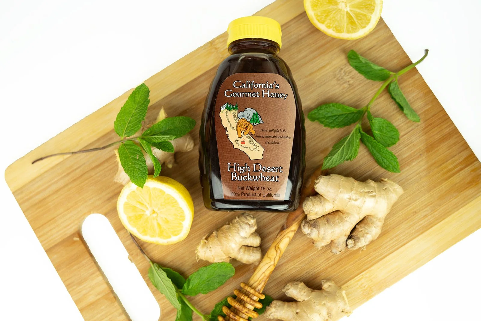 A jar of California's Gourmet Honey high desert buckwheat honey on a wooden cutting board surrounded by lemon slices, fresh mint leaves, ginger, and a honey dipper.