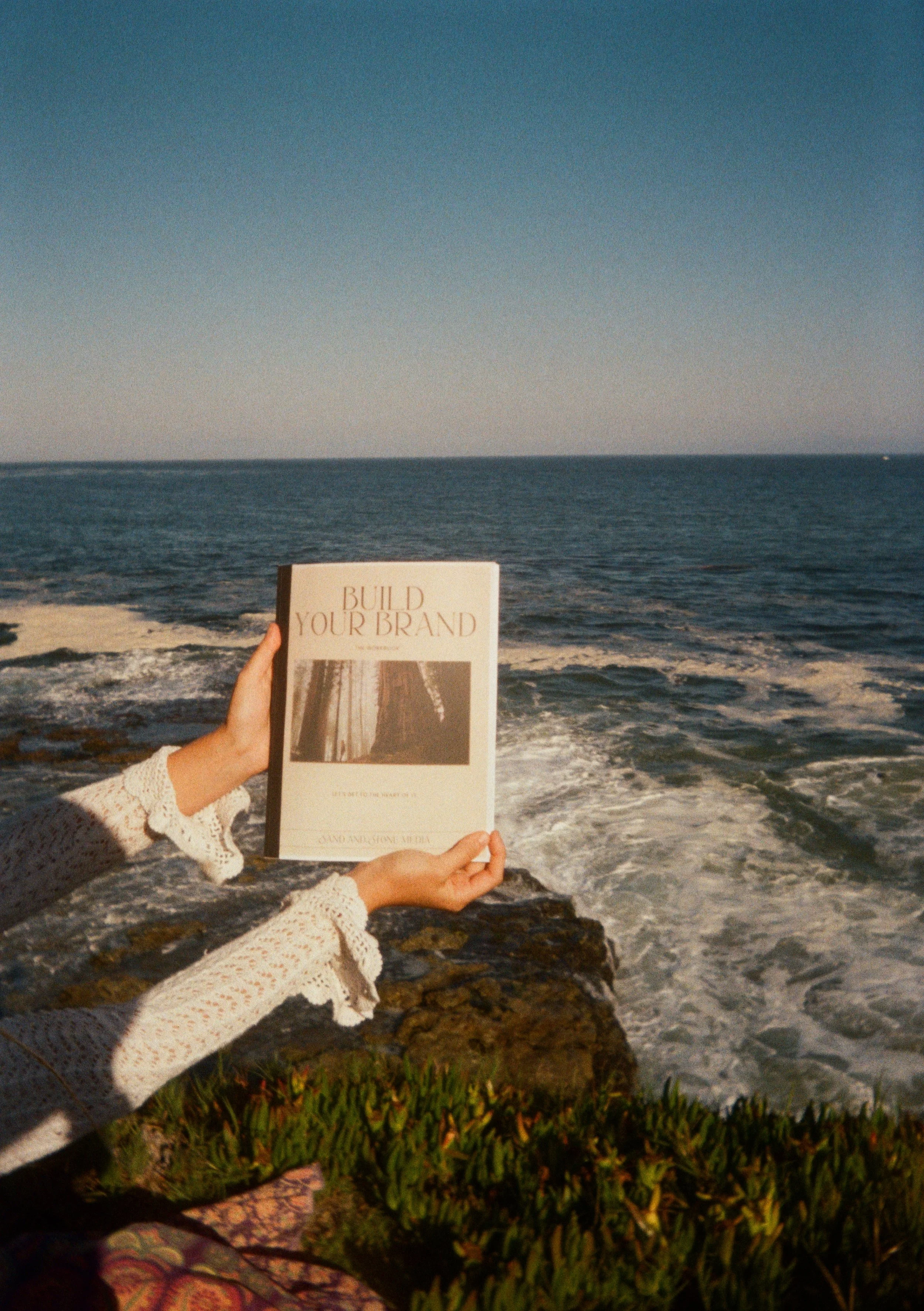 A person holds a book titled 'Build Your Brand' in front of the ocean at sunset, with waves crashing on rocks and greenery in the foreground.