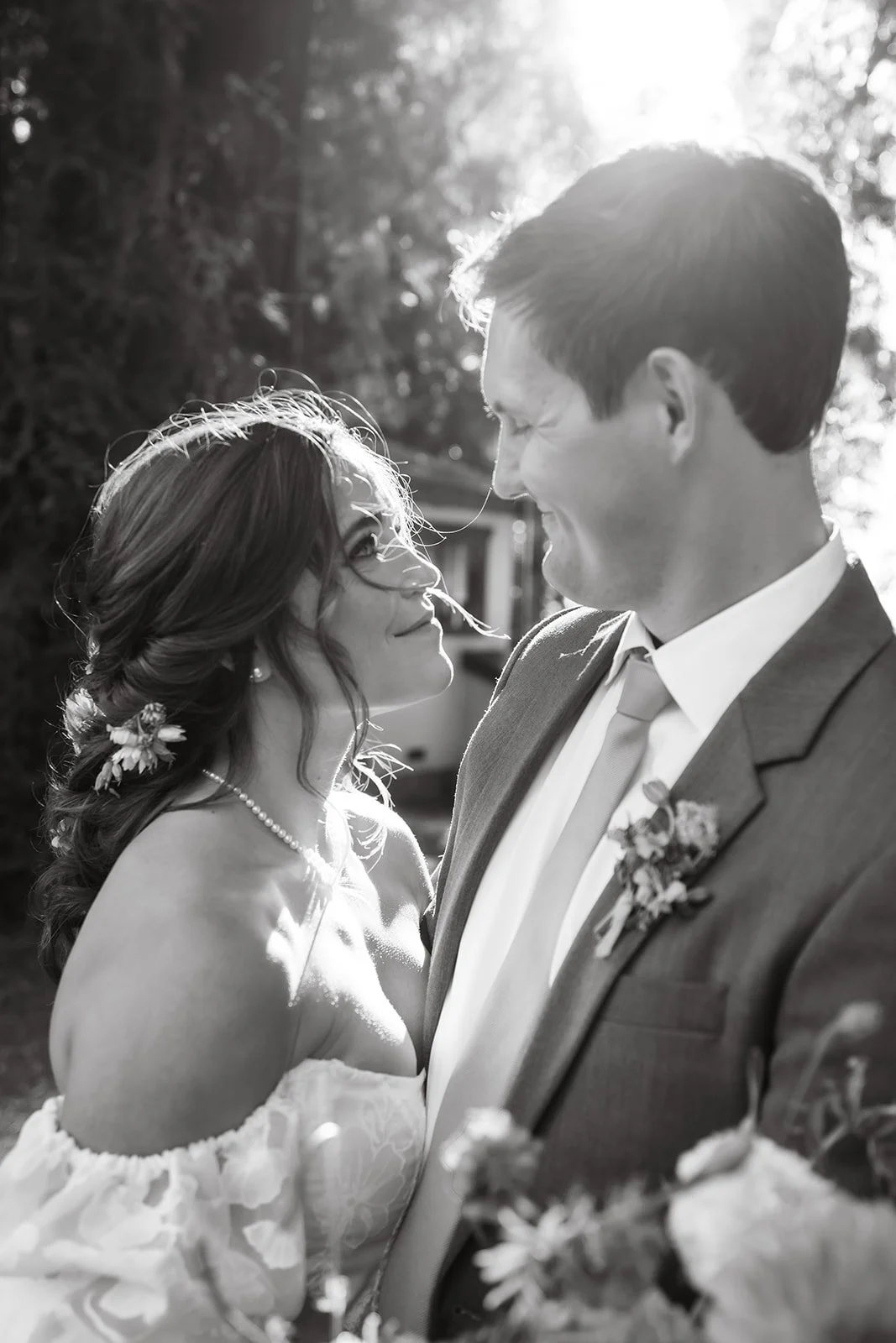 A black-and-white photograph of a bride and groom smiling at each other outdoors during sunset, with the sun shining behind them.