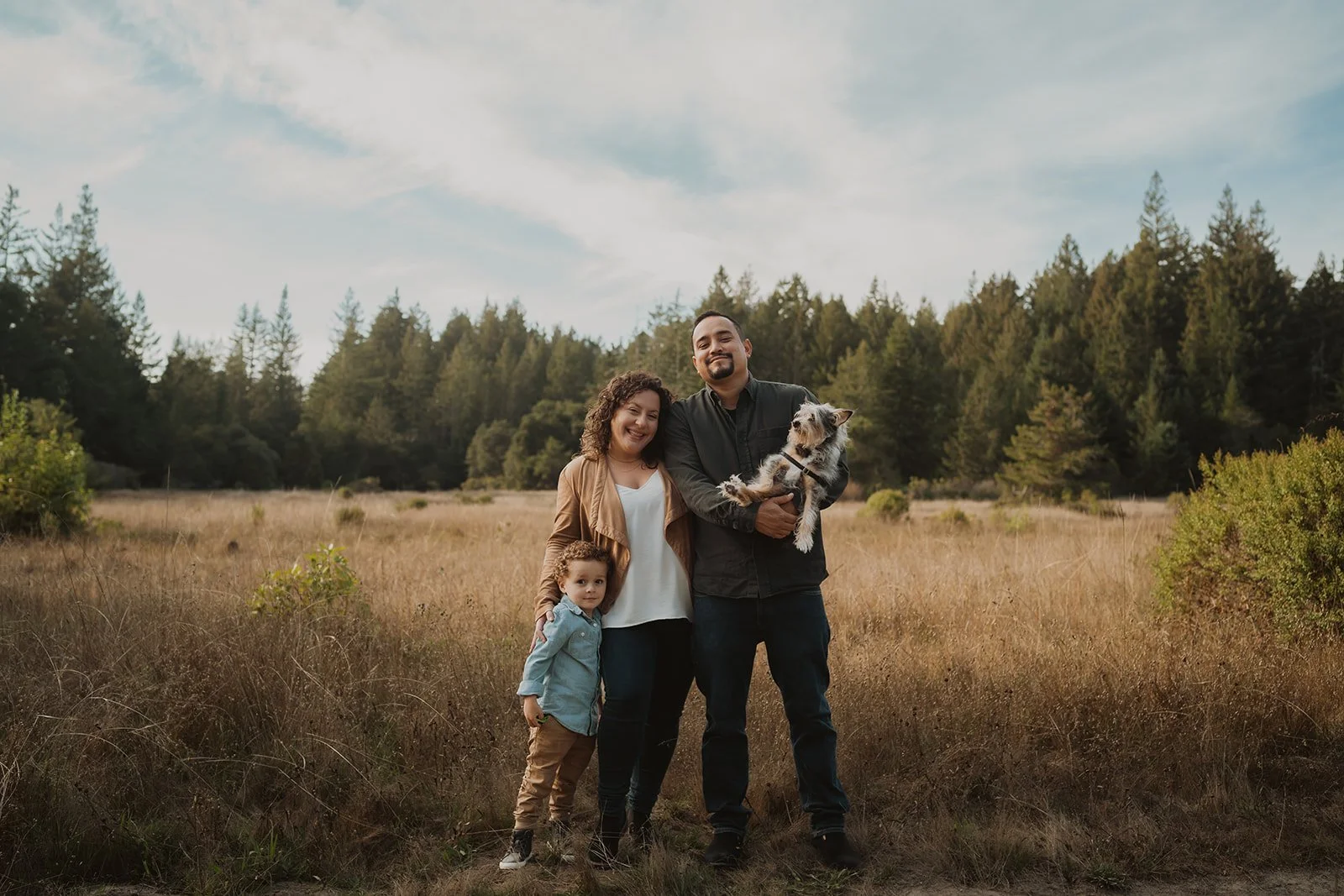 A family of four with a dog standing in a grassy field with trees in the background and a partly cloudy sky.