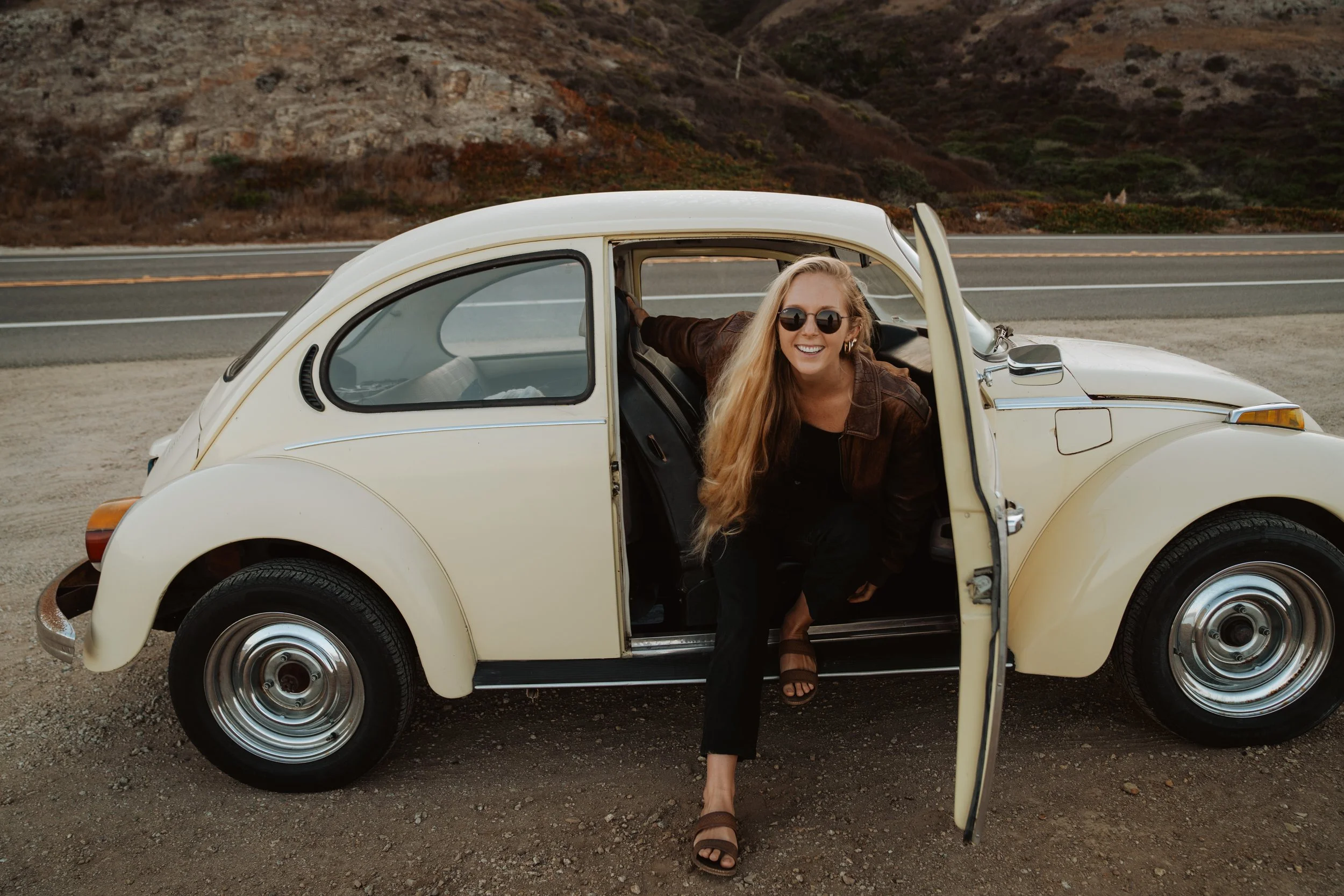 A woman with long blonde hair, wearing sunglasses, a brown jacket, and sandals, smiling and getting out of a cream-colored vintage Volkswagen Beetle parked by a roadside on a scenic route with hills in the background.