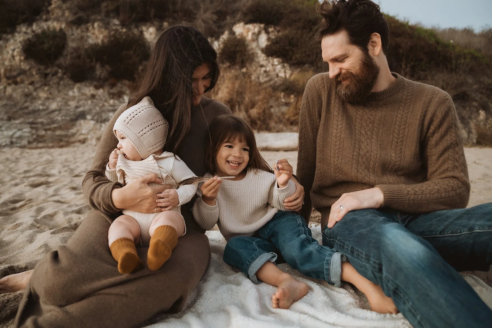 A family of four sitting on a sandy beach, smiling and enjoying each other's company. The mother is holding a small child, and the father is sitting nearby, with a young girl sitting in front of him. The family is dressed in casual, warm clothing.