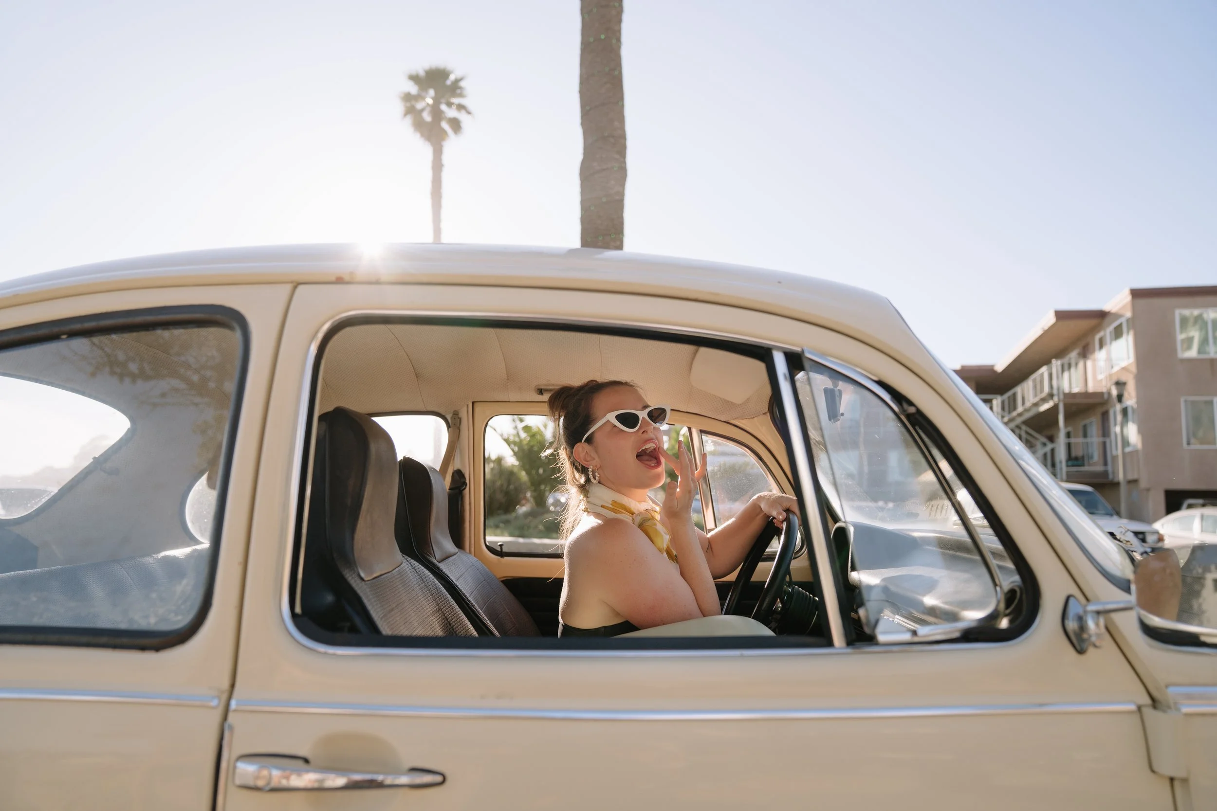 A woman with sunglasses sitting in the driver's seat of a vintage beige car, singing and gesturing with her left hand, with palm trees and apartment buildings in the background.