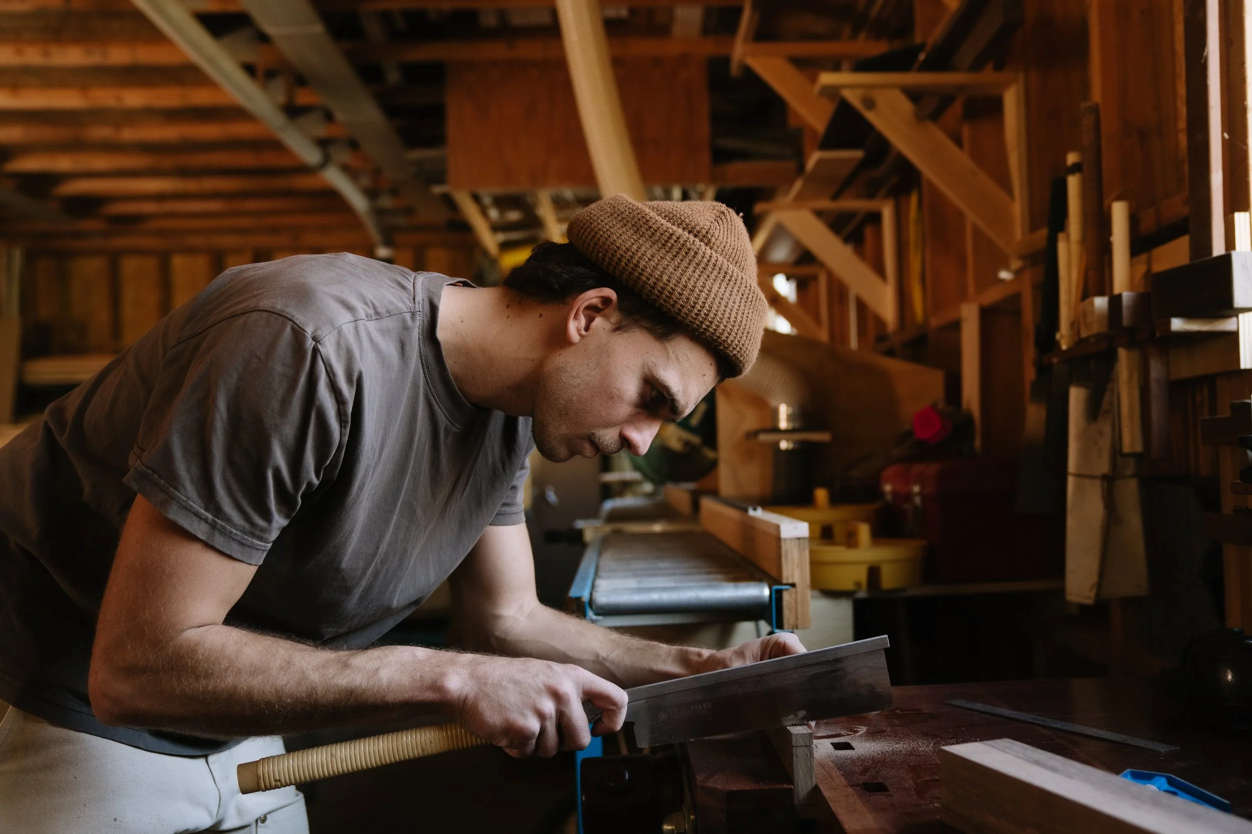 A man sanding a piece of wood at a woodworking shop, wearing a brown beanie and a gray t-shirt.