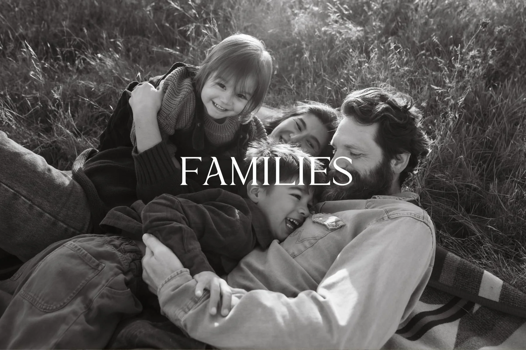 A black and white photo of a family, including an adult man, an adult woman, and three children, lying on grass outdoors, smiling and embracing each other. The word "FAMILIES" is overlaid in the center.