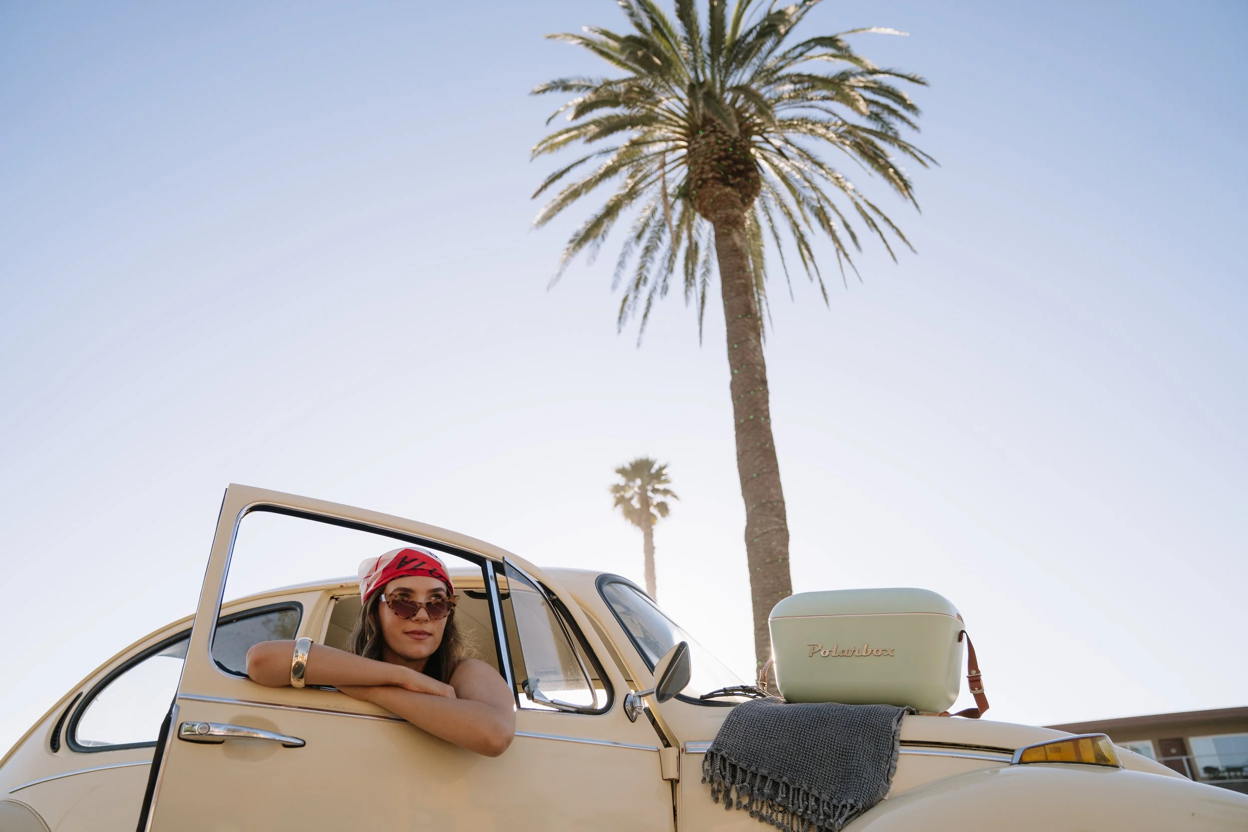 A woman rests her arm on the open window of a cream-colored vintage Volkswagen Beetle parked outdoors under tall palm trees, with a clear sky in the background. She wears sunglasses, a scarf, and a bracelet.